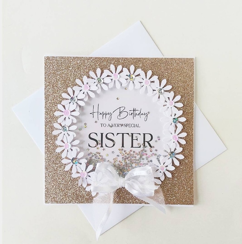 May include: A gold glitter card with a white daisy wreath and a white bow. The card says "Happy Birthday to a very special Sister".