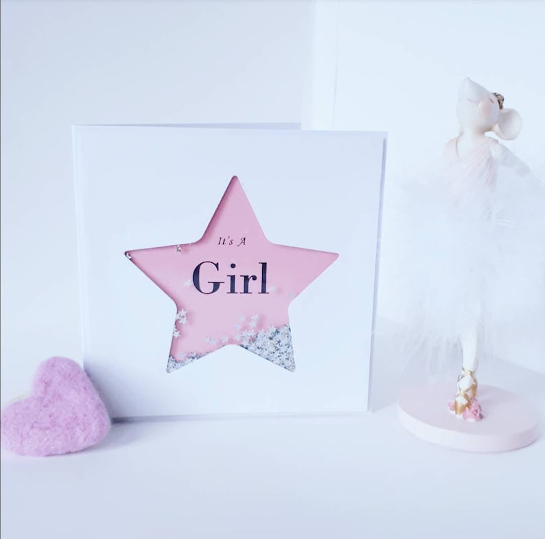 K&ouml;nnte beinhalten: Eine rosa und wei&szlig;e Karte mit einem sternf&ouml;rmigen Ausschnitt. Der Stern ist mit rosa Papier und silbernem Glitzer gef&uuml;llt. Der Text "It's a Girl" ist in Schwarz in den Stern gedruckt.