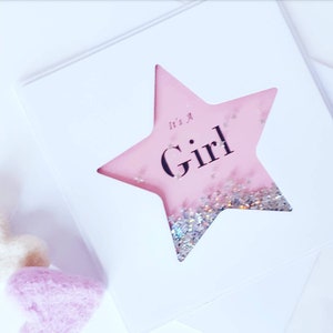 K&ouml;nnte beinhalten: Eine rosa sternf&ouml;rmige Karte mit Glitzer im Inneren. Die Karte tr&auml;gt den Text "It's a Girl".