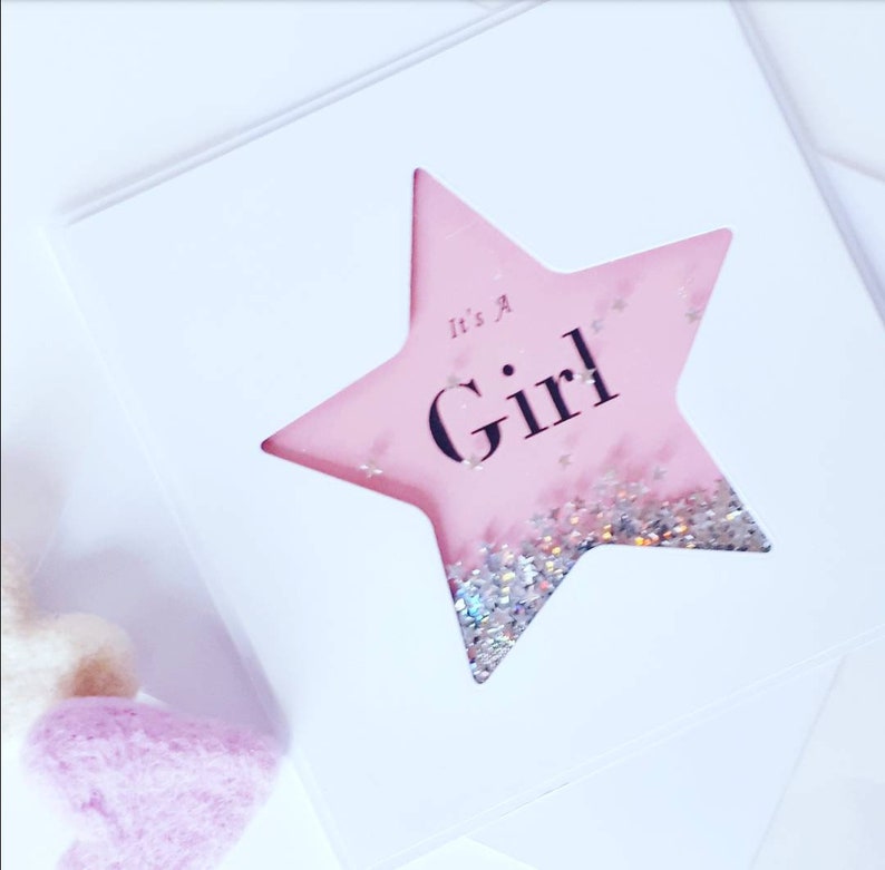 K&ouml;nnte beinhalten: Eine rosa sternf&ouml;rmige Karte mit Glitzer im Inneren. Die Karte tr&auml;gt den Text "It's a Girl".