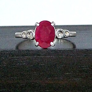 May include: A silver ring with a large, oval-cut ruby gemstone as the centerpiece. The ruby is a deep red color and is held in place by four prongs. The ring has small, circular accents on either side of the ruby.