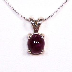 May include: A silver pendant necklace featuring a smooth, oval-shaped deep red gemstone set in a four-prong metal mounting. The pendant is suspended from a delicate, thin silver chain that extends upwards from the top of the setting.