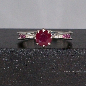 May include: A silver ring with a large red ruby gemstone surrounded by smaller ruby stones. The ring has a delicate, curved design.