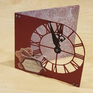 May include: Red and white paper card with a cut-out clock face. The clock hands are set to midnight. The card has a small tag attached with the text "Merry Christmas and a Happy New Year".