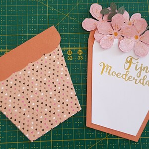 Peut inclure: Une carte faite à la main avec un motif de pot de fleurs. Le pot est en carton orange et a un motif de cœurs roses et bruns. La carte est décorée de fleurs en papier rose et une inscription dorée qui dit "Fijne Moederdag", ce qui se traduit par "Joyeuse fête des mères" en français.