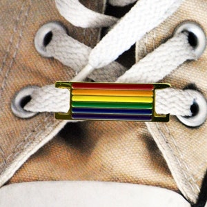 May include: A pair of white canvas shoes with a rainbow-colored shoelace charm. The charm is a metal bar with six stripes of red, orange, yellow, green, blue, and purple.