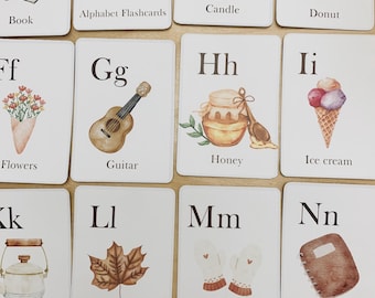 May include: Alphabet flashcards with illustrations of various objects, such as a book, candle, donut, flowers, guitar, honey, ice cream, kettle, leaf, mittens, notebook. The flashcards are white with brown lettering and illustrations.