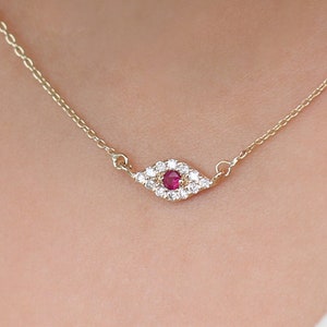 May include: A gold chain necklace with a small evil eye pendant. The pendant is made of white stones with a single red stone in the center.