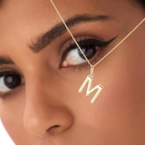 May include: A gold necklace featuring the letter "M" as a pendant. The pendant is a capital letter with a three-dimensional design. The necklace is worn by a person with dark hair and skin, against a white backdrop.