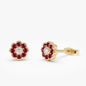 Könnte beinhalten: Ein Paar goldene Ohrringe in Blumenform mit einem einzelnen weißen Diamanten in der Mitte jeder Blume und umgeben von roten Edelsteinen.