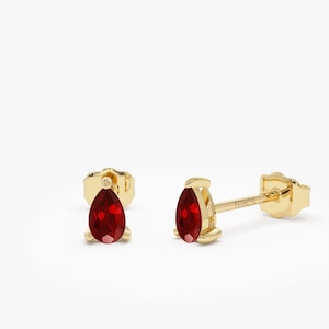 May include: A pair of gold earrings with pear-shaped red gemstones. The earrings have a simple design and are perfect for everyday wear.
