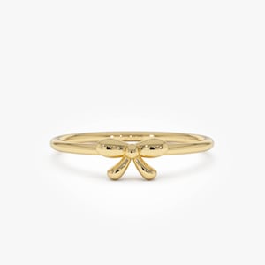 May include: A delicate gold ring featuring a bow design. The ring has a thin band and a small, three-dimensional bow at the center. The bow has rounded edges and a polished finish, reflecting light. The ring is set against a clean white background.