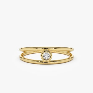 May include: A gold ring with a single round diamond set in a bezel setting. The ring is made of two bands that are stacked on top of each other.