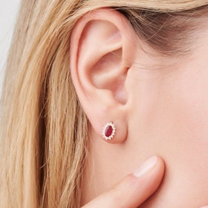 May include: A pair of gold stud earrings featuring a red oval gemstone surrounded by smaller white gemstones.