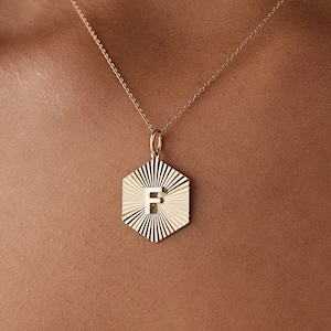 May include: A gold necklace with a hexagonal pendant featuring a sunburst design and the letter "F" in the centre.