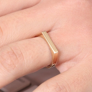 May include: A gold-colored ring with a square band. The ring is worn on a finger.
