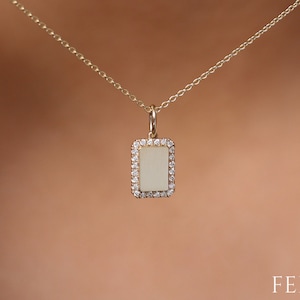 May include: A gold necklace featuring a rectangular pendant with a light-coloured centre, surrounded by small, sparkling stones. The necklace is shown against a neutral background.