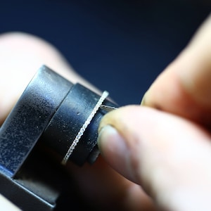 May include: A close-up of a ring being sized with a metal sizing tool. The ring has a thin band with small diamonds set in a pave setting.
