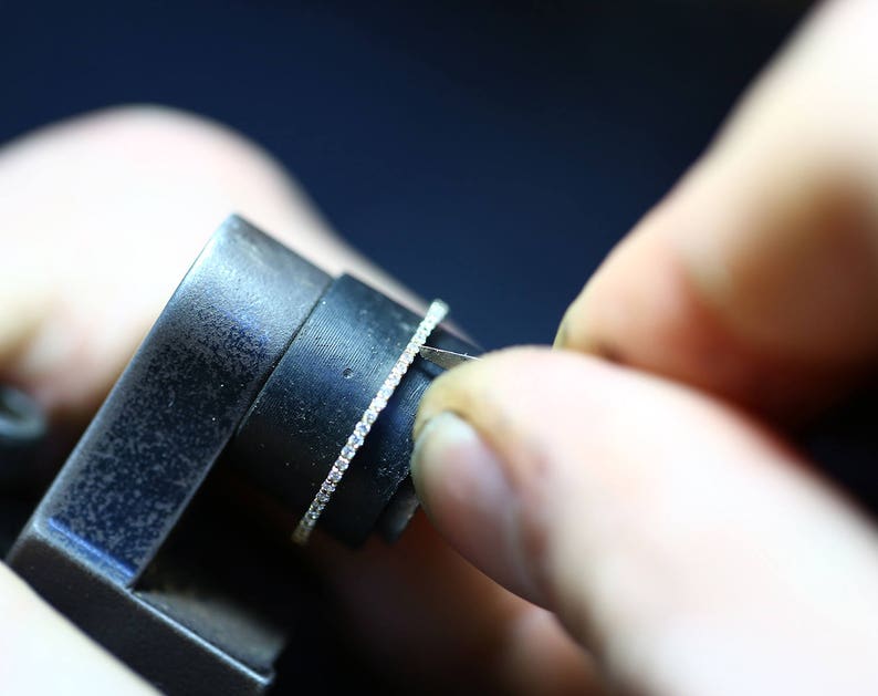 May include: A close-up of a ring being sized with a metal sizing tool. The ring has a thin band with small diamonds set in a pave setting.