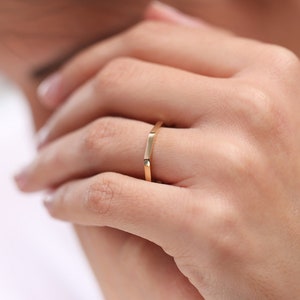 May include: A simple gold band ring with a slightly squared shape. The ring is worn on the ring finger of a person's left hand.