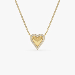 May include: A gold-colored heart pendant necklace. The heart-shaped pendant has a solid gold center, surrounded by a halo of small, clear gemstones. The necklace chain is also gold-colored and delicate, with a simple link design.