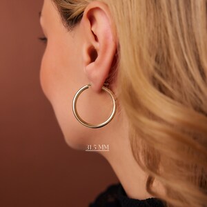 Puede incluir: Pendientes de aro de oro con un di&aacute;metro de 31,5 mm. Los pendientes se muestran en la oreja de una persona.