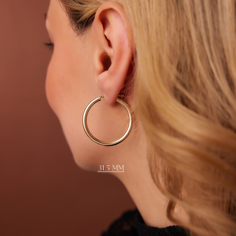 Puede incluir: Pendientes de aro de oro con un di&aacute;metro de 31,5 mm. Los pendientes se muestran en la oreja de una persona.