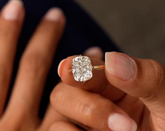 May include: A gold ring featuring a large, square-cut diamond. The diamond is clear and sparkling, set on a thin gold band. The ring is held up, showcasing its design and brilliance. A classic piece of jewelry.
