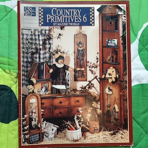 May include: A softcover book titled Country Primitives 6 by Maxine Thomas. The cover features a photograph of a wooden cabinet, a dresser, and various decorative items including a bear figurine, birdhouses, and floral arrangements.