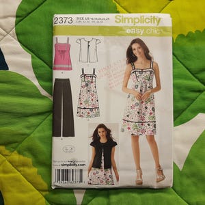 May include: A Simplicity sewing pattern, number 2373, featuring a woman in a floral dress. The pattern includes designs for a top, jacket, trousers, and a dress. The cover is set against a green and white patterned background.