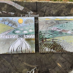 May include: Two ceramic tiles with a countryside scene. The tile on the left has a yellow sun, green hills, and white sheep. The tile on the right has a green hillside with houses and a brown fence.
