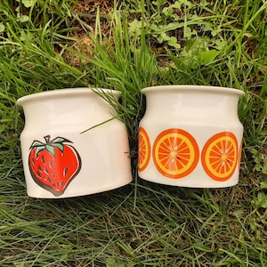 May include: Two white ceramic jars with colourful fruit designs. One jar has a red strawberry design and the other has three orange slices.