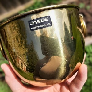 May include: A gold-coloured metal planter with a black label that reads "100% Messing Made in Denmark".