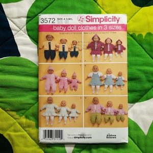 May include: Simplicity baby doll clothes pattern in three sizes, featuring various outfits for dolls. The pattern includes options for jackets, dresses, and overalls. The pattern is on a white background with a green and white patterned fabric.