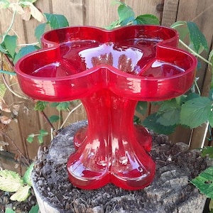 May include: A red glass vase with a four-leaf clover design. The vase has a wide, shallow base and a tall, slender neck.