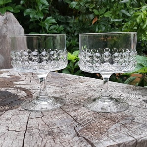 May include: Two clear glass stemmed dessert dishes with a textured bubble design. The dishes are sitting on a wooden surface.