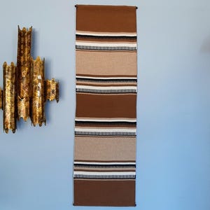 May include: A long, rectangular woven wall hanging with horizontal stripes in shades of brown, tan, black, and white. The textile is displayed vertically against a light blue wall. The wall hanging is a decorative piece.