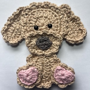 May include: A crocheted dog applique in beige yarn with black eyes and a grey nose. The dog's paws are pink. This handmade item is suitable for various craft projects, such as embellishing clothing or accessories.