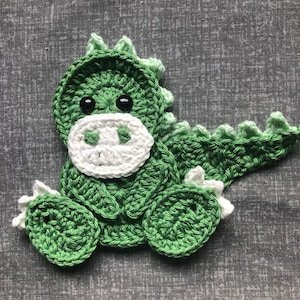 May include: A green crocheted dinosaur with white eyes and a white snout. The dinosaur is sitting on a grey fabric background.