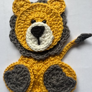 May include: A crocheted lion applique in yellow, grey, white, and black. The lion has a yellow face and body, a grey mane and paws, a white muzzle, and a black nose and eyes. The tail is yellow and grey. This handmade item is suitable for various craft projects.