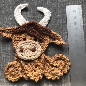 May include: A crocheted Highland cow applique in shades of brown, tan, and white. The cow has white horns, a tan muzzle, and dark eyes. A ruler is placed next to the cow, showing measurements in millimeters and centimeters.