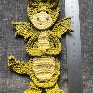 May include: A crocheted dragon toy in olive green and pale yellow. The dragon features a detailed head, wings, and a textured body. A ruler is placed beside the dragon, indicating its size. The dragon's height is approximately 19 cm.