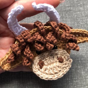 May include: A crocheted brown and white highland cow ornament with white yarn horns.