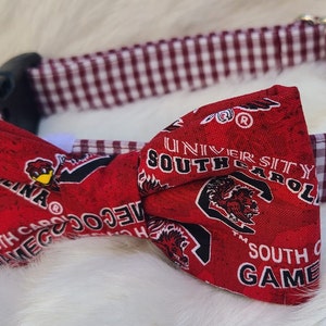 May include: A red and white dog collar with a red bow tie featuring the University of South Carolina Gamecocks logo. The bow tie has a distressed look and the collar is a gingham pattern.