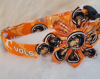 Könnte beinhalten: Oranges Hundehalsband mit Blumenakzent. Das Halsband hat ein sich wiederholendes Muster eines Hundegesichts und das Wort "VOLS". Der Blumenakzent ist aus dem gleichen Stoff gefertigt und hat eine schwarze Schnalle und einen silbernen Metallring.