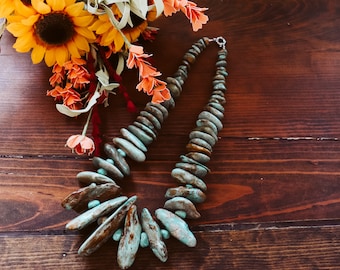 May include: A turquoise and brown stone necklace with a silver clasp. The necklace is made of irregularly shaped stones and is displayed on a wooden surface. A bouquet of sunflowers and orange flowers is in the background.