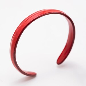 Puede incluir: Un brazalete de puño rojo brillante. El brazalete abierto tiene un diseño suave y redondeado. El color rojo es consistente en todo el brazalete, que parece estar hecho de un material sólido. El brazalete está sobre un fondo blanco.