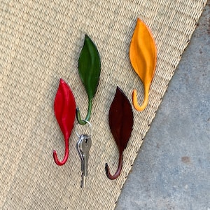 May include: Four leather leaf-shaped hooks in red, green, brown, and yellow, mounted on a woven mat. A silver key is hanging on the red hook.