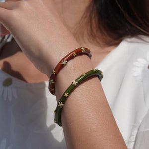 May include: Two leather bracelets with embroidered flower designs. One bracelet is brown with yellow flowers and the other is green with white flowers.