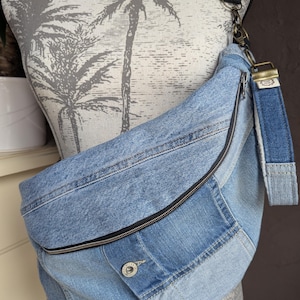 Peut inclure: Un sac bandoulière en jean bleu clair avec une fermeture éclair noire et une sangle noire. Le sac est doté d'une fermeture à bouton et d'une sangle en jean. Le sac est présenté sur un mannequin avec un motif de palmier.
